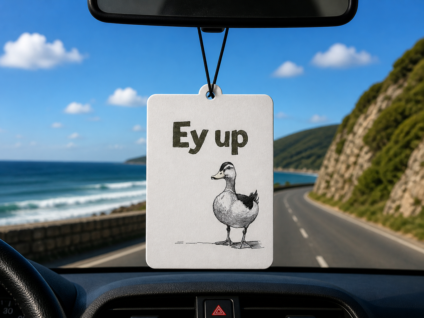 Car air freshener with a duck illustration and 'Ey up' text hanging from a car's rearview mirror with a scenic road and ocean view.