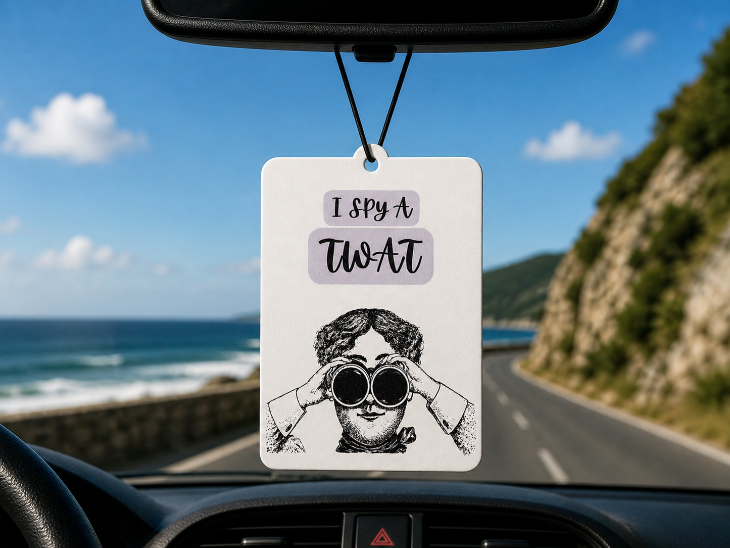 Car air freshener with humorous design hanging from a car's rearview mirror, with a scenic road and ocean view outside.