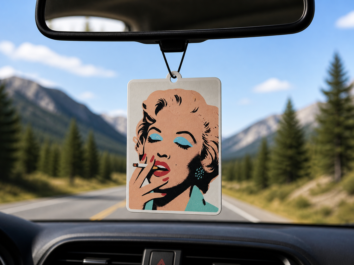Car air freshener with a stylized face design hanging from a car's rearview mirror with a scenic road in the background.