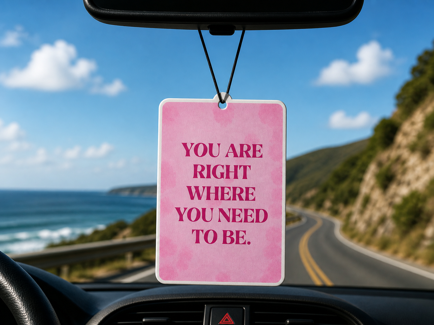 Pink car air freshener with motivational quote hanging from a car's rearview mirror with scenic road and ocean view.