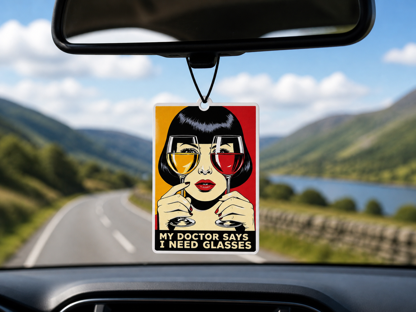 Car air freshener with a graphic design of a woman holding glasses, hanging from a car's rearview mirror with a scenic road in the background.
