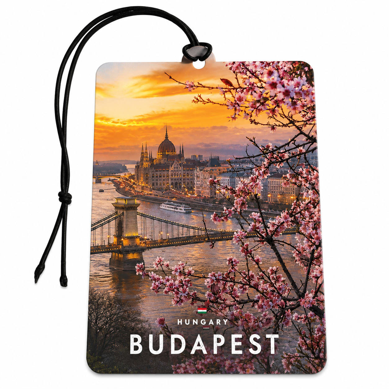 A rectangular Budapest Car Air Freshener or luggage tag featuring a scenic sunset cityscape with the Hungarian Parliament Building, Chain Bridge, and pink blossoms in the foreground. The word BUDAPEST appears at the bottom, with a small Hungary flag above it.