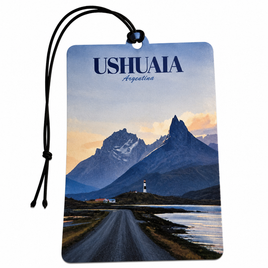 A rectangular luggage tag or car air freshener tag with a black hanging cord, printed with a scenic landscape of Ushuaia, Argentina. The design features a road leading toward a lighthouse between dark blue mountains under a pale sky with warm sunset clouds.