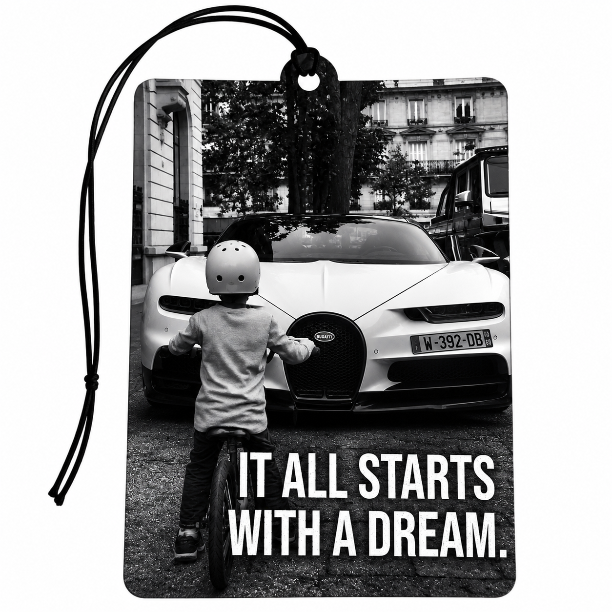 A rectangular printed car air freshener or tag with a black hanging loop, featuring a black-and-white photo of a child in a helmet looking at a white Bugatti car on a street. Large white text at the bottom reads, 'IT ALL STARTS WITH A DREAM.