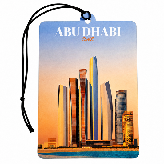 A rectangular Abu Dhabi car air freshener tag featuring a printed skyline of the city with the text 'ABU DHABI uae' at the top. The tag has a blue sky gradient background, a warm orange sunset over the skyscrapers, and a black hanging cord, shown on a plain white background.