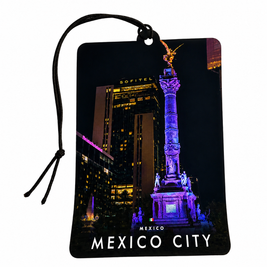 A rectangular car air freshener featuring a nighttime photograph of Mexico City with the Monumento a la Independencia illuminated in purple, set against a black city skyline. The tag has a black hanging loop and the words 'MEXICO CITY' printed in white at the bottom.